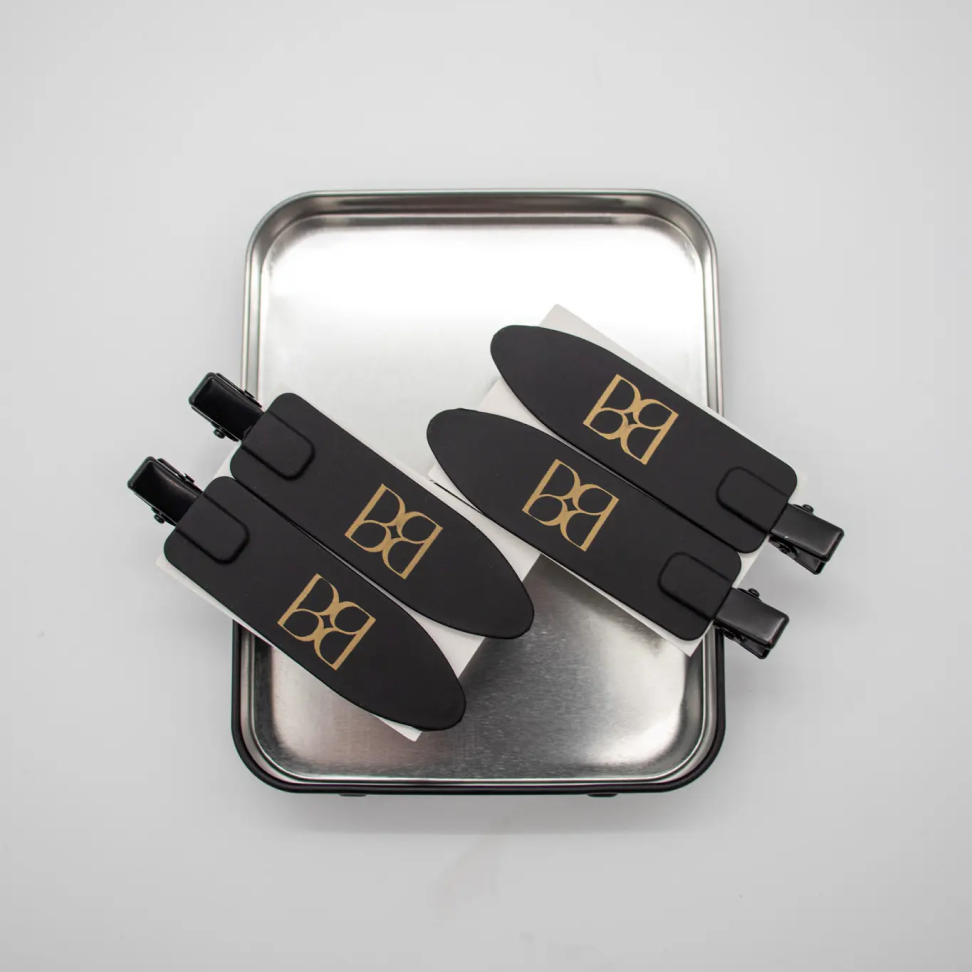 Black hair clips with gold 'BB' logo on a metallic tray against a light gray background